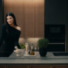 Laura Pausini nella cucina Poetica con ante telaio in Impiallacciato Frassino Cacao e ante Pet in Microdecor Space Black; a destra boiserie Vertical System. Sullo sfondo, la composizione con pensili Sail completa l’ambiente